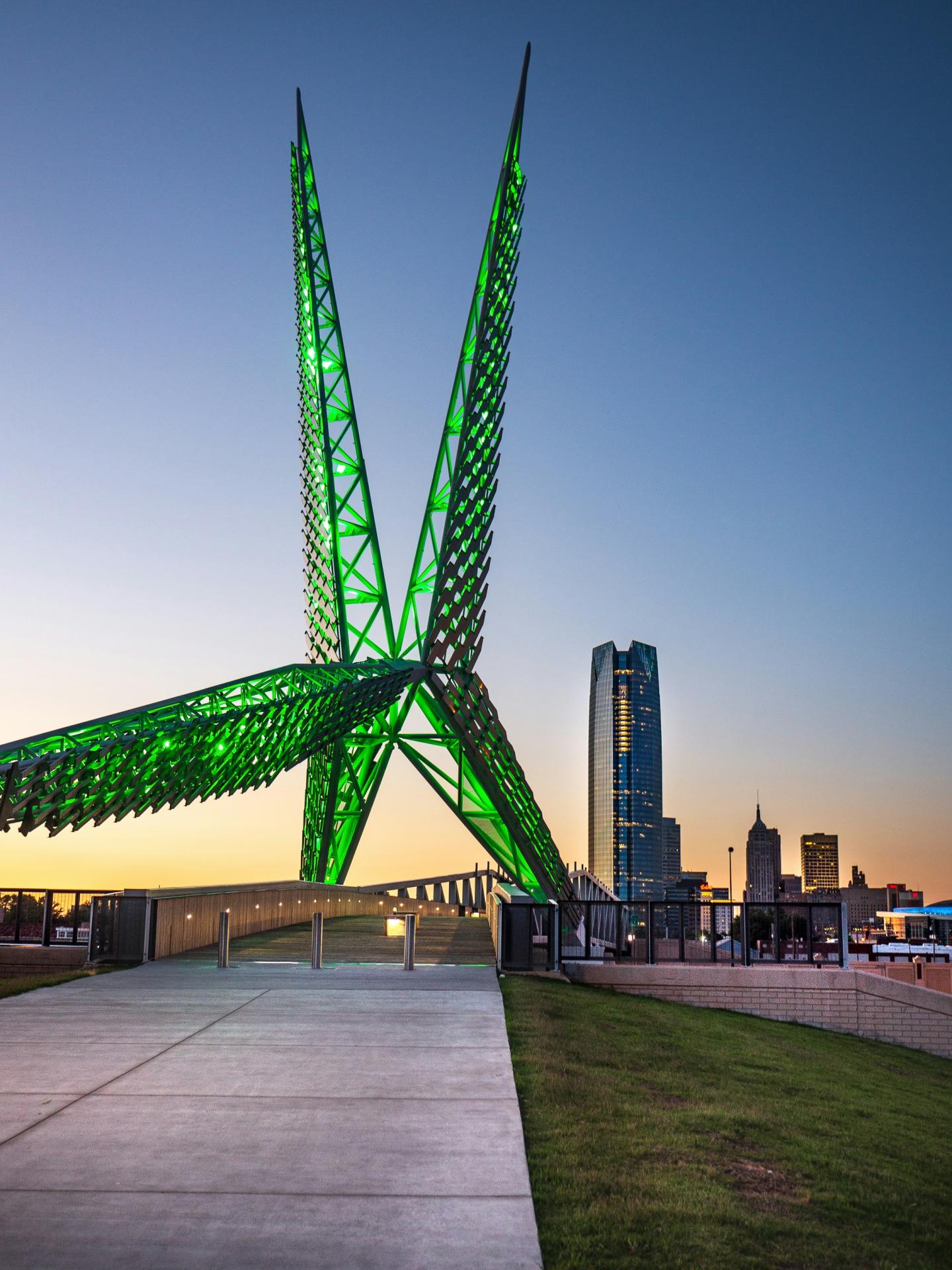 Green-lit sculpture with city skyline at dusk.