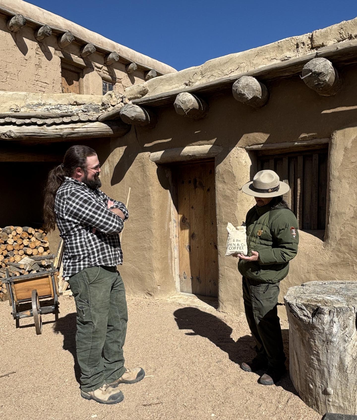 Two people converse outside an adobe building under clear blue sky.