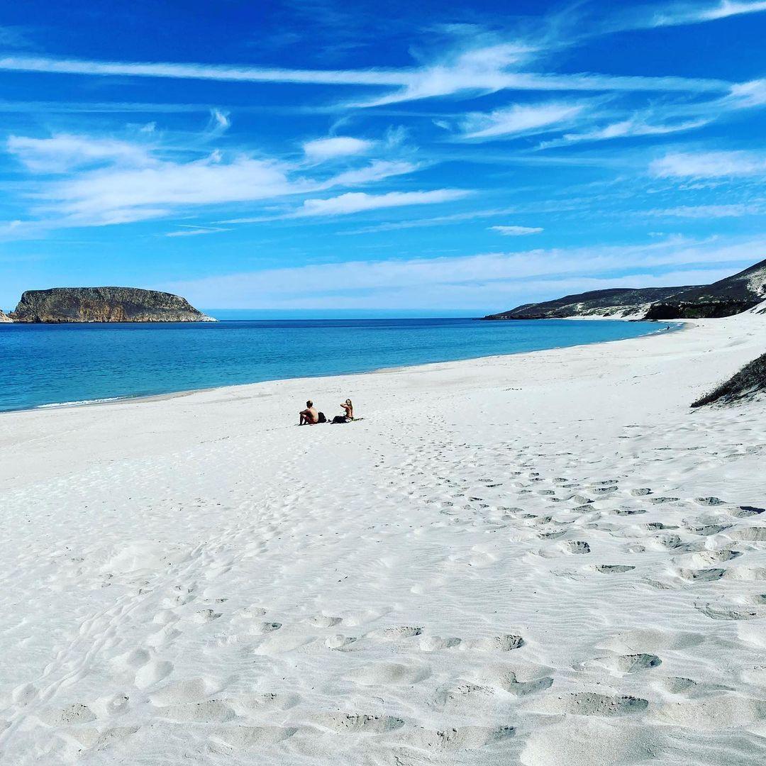 White sandy beach with two people, blue sky, and calm sea.