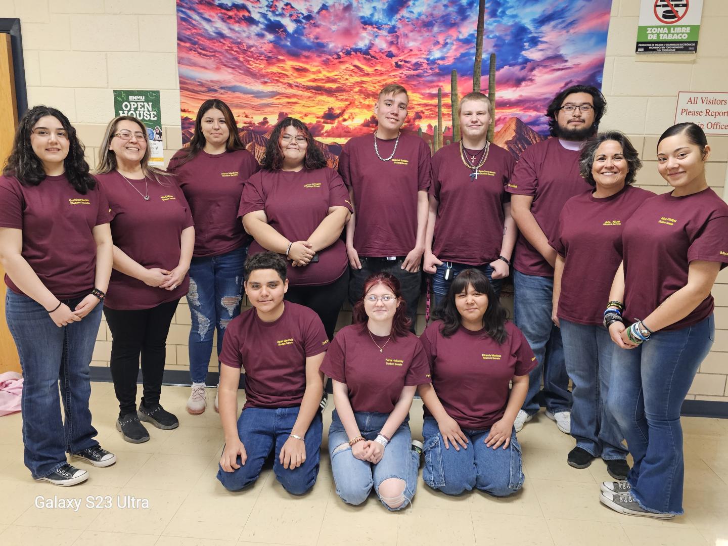 Group of people in matching maroon shirts, in front of a colorful sunset painting indoors.