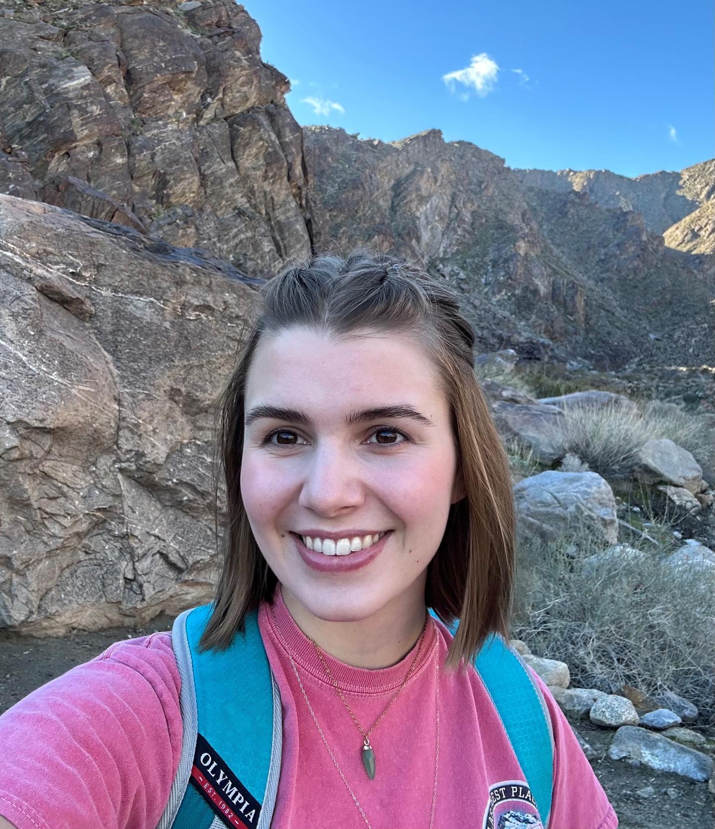 Young woman smiling on a rocky mountain trail, wearing a pink shirt and turquoise backpack.