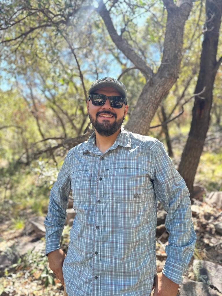 Man in plaid shirt and sunglasses smiles in a sunny forest setting.