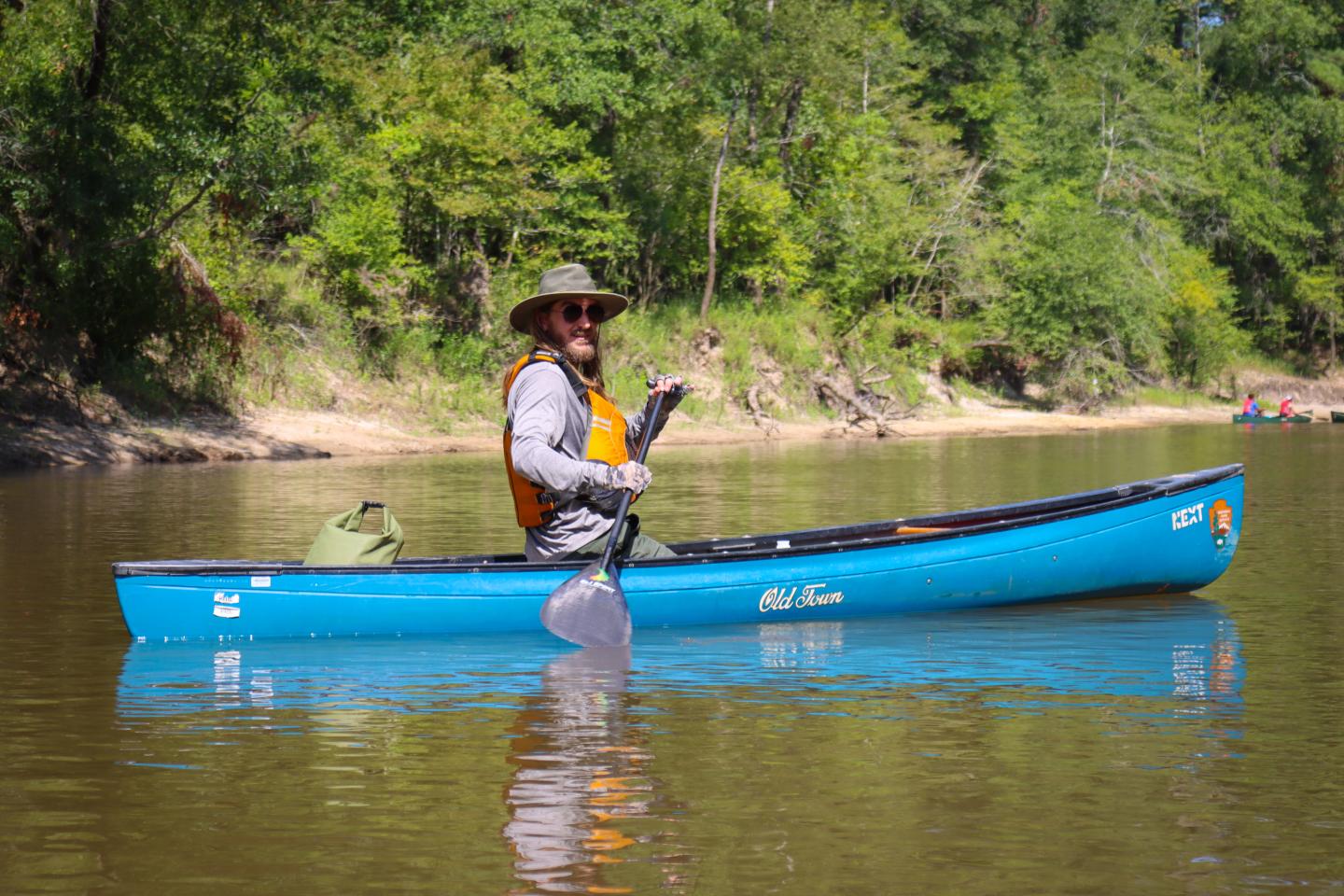 Man paddling blue canoe on calm river, surrounded by trees.