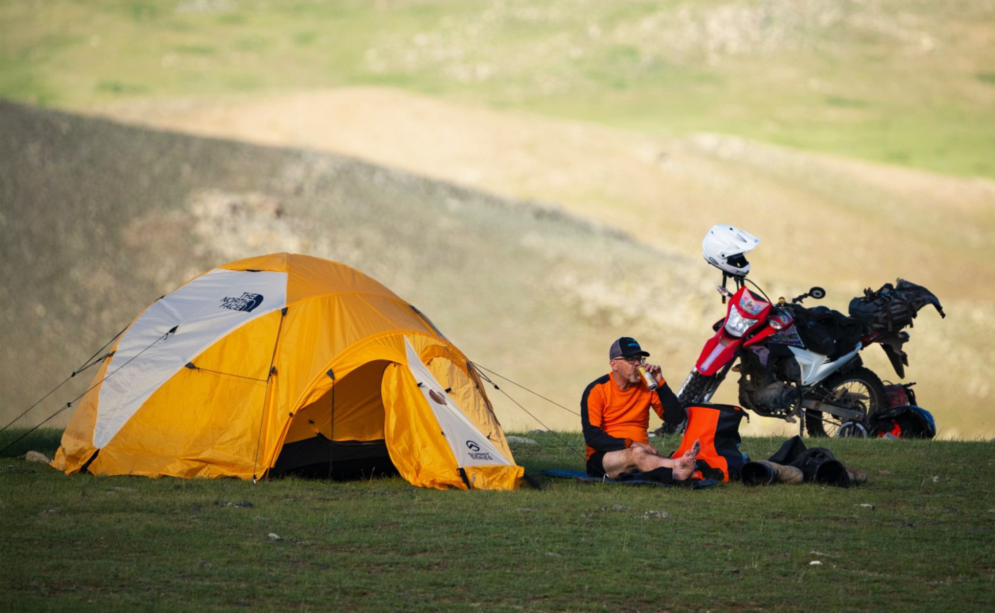 Orange tent and motorcycle on grassy hillside with person resting nearby.