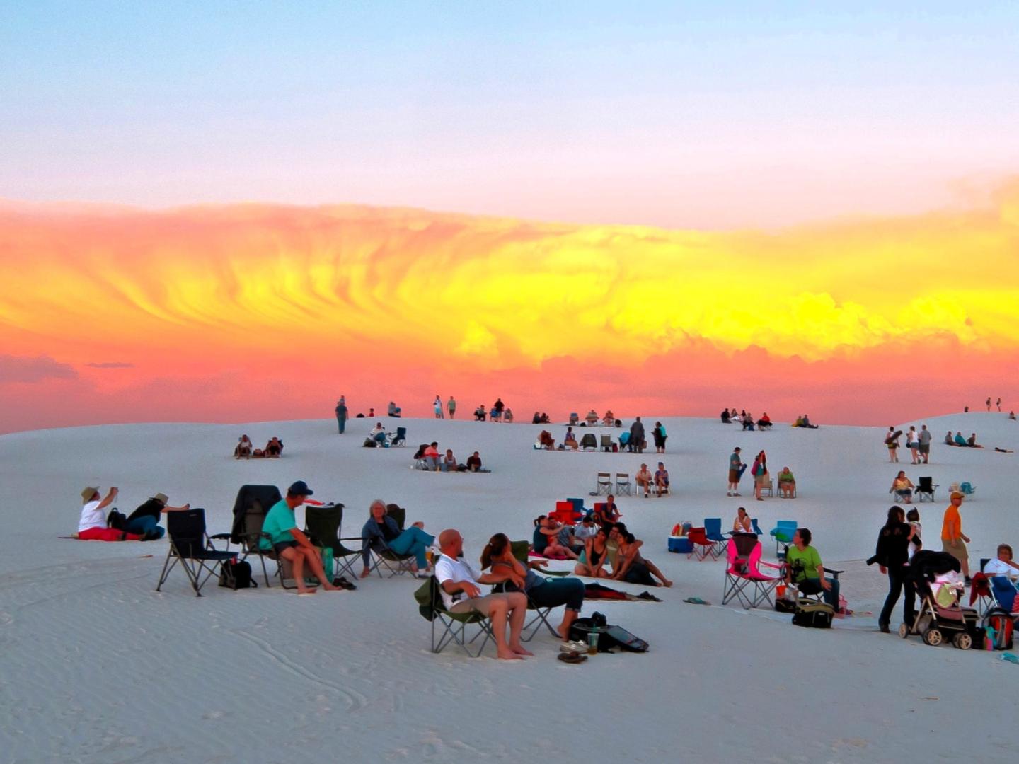 People seated on white sand dunes at sunset, sky glowing in orange and pink hues.
