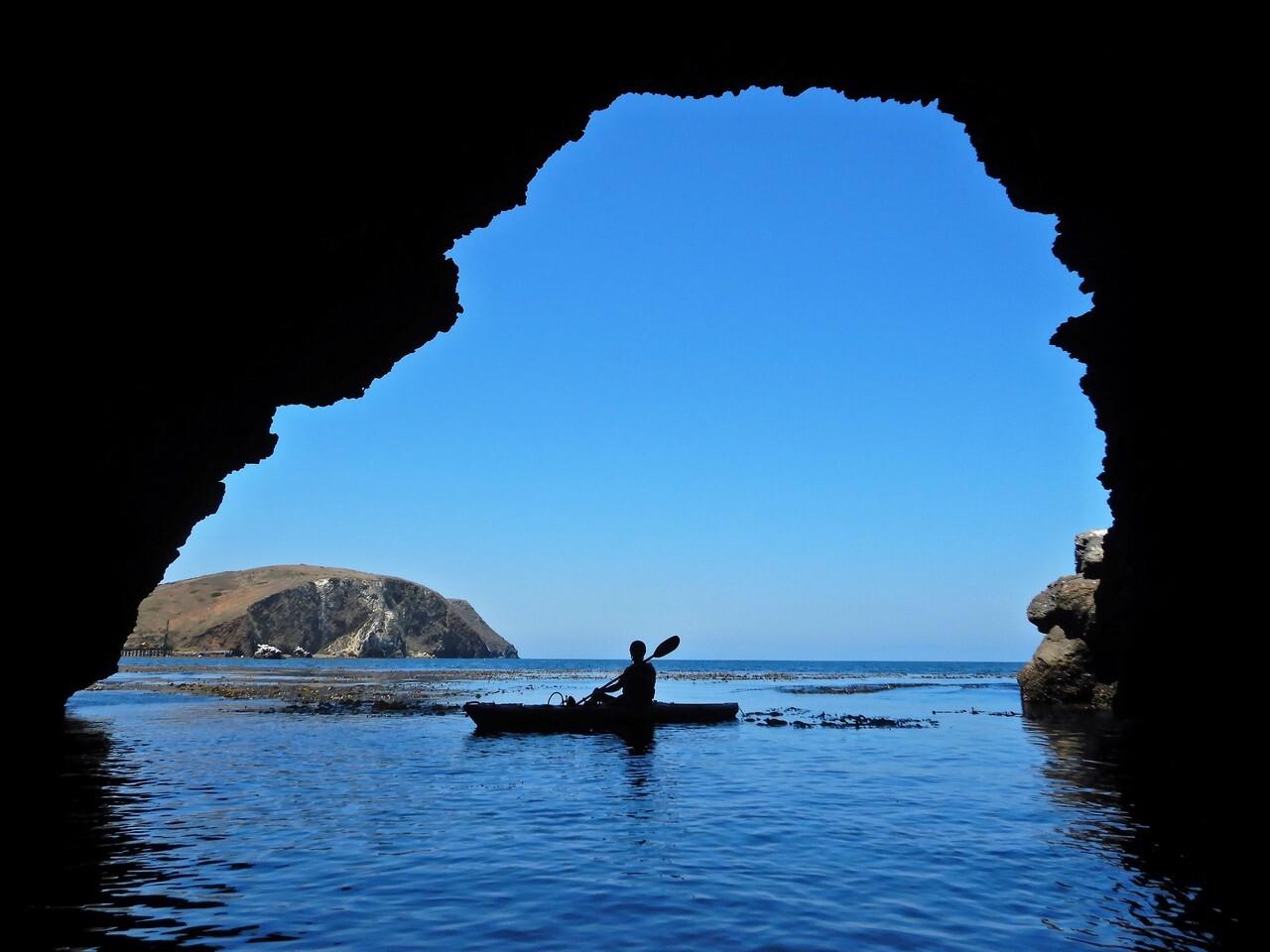 Kayaker silhouetted in a cave, vibrant blue sky and ocean outside.