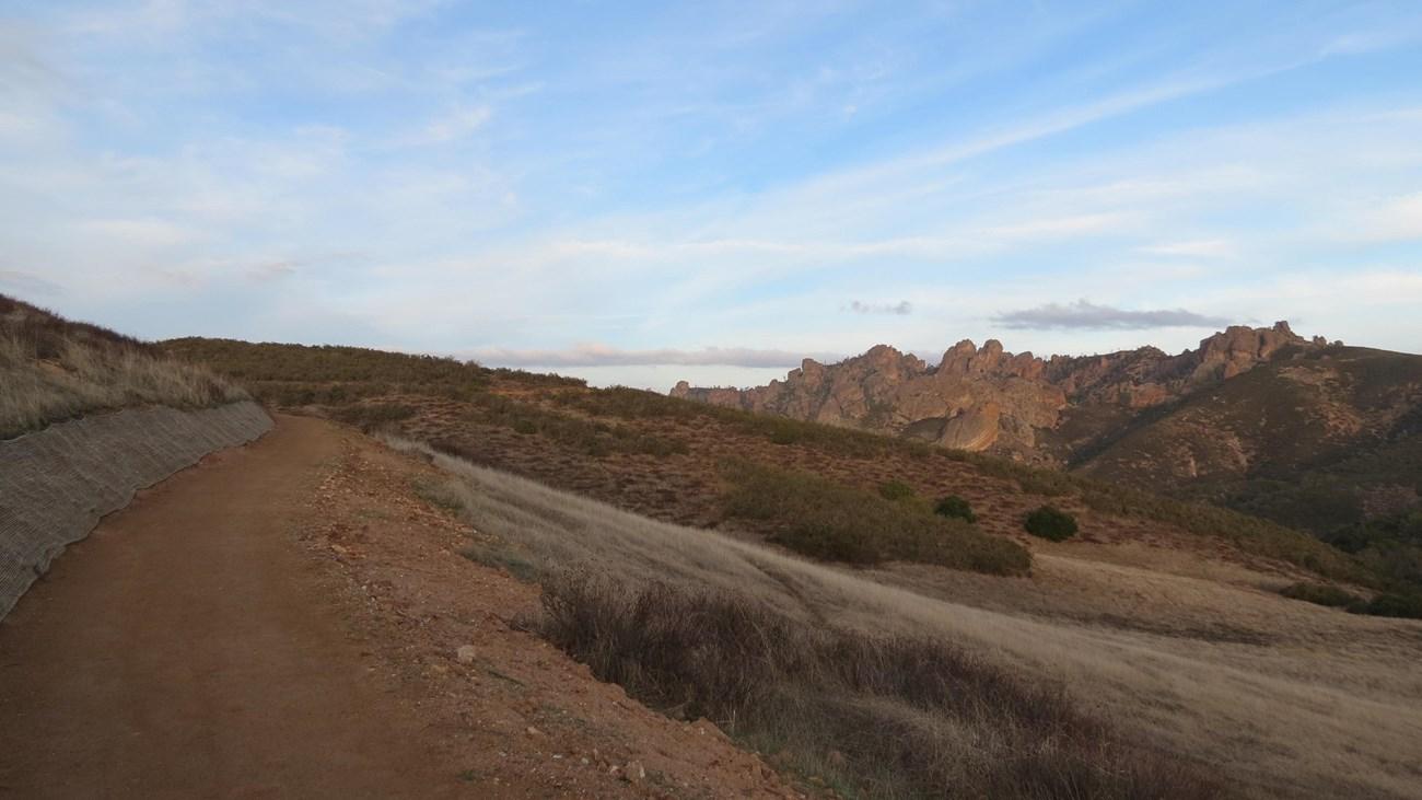 Dirt path through dry hills with distant rocky mountains under blue sky.