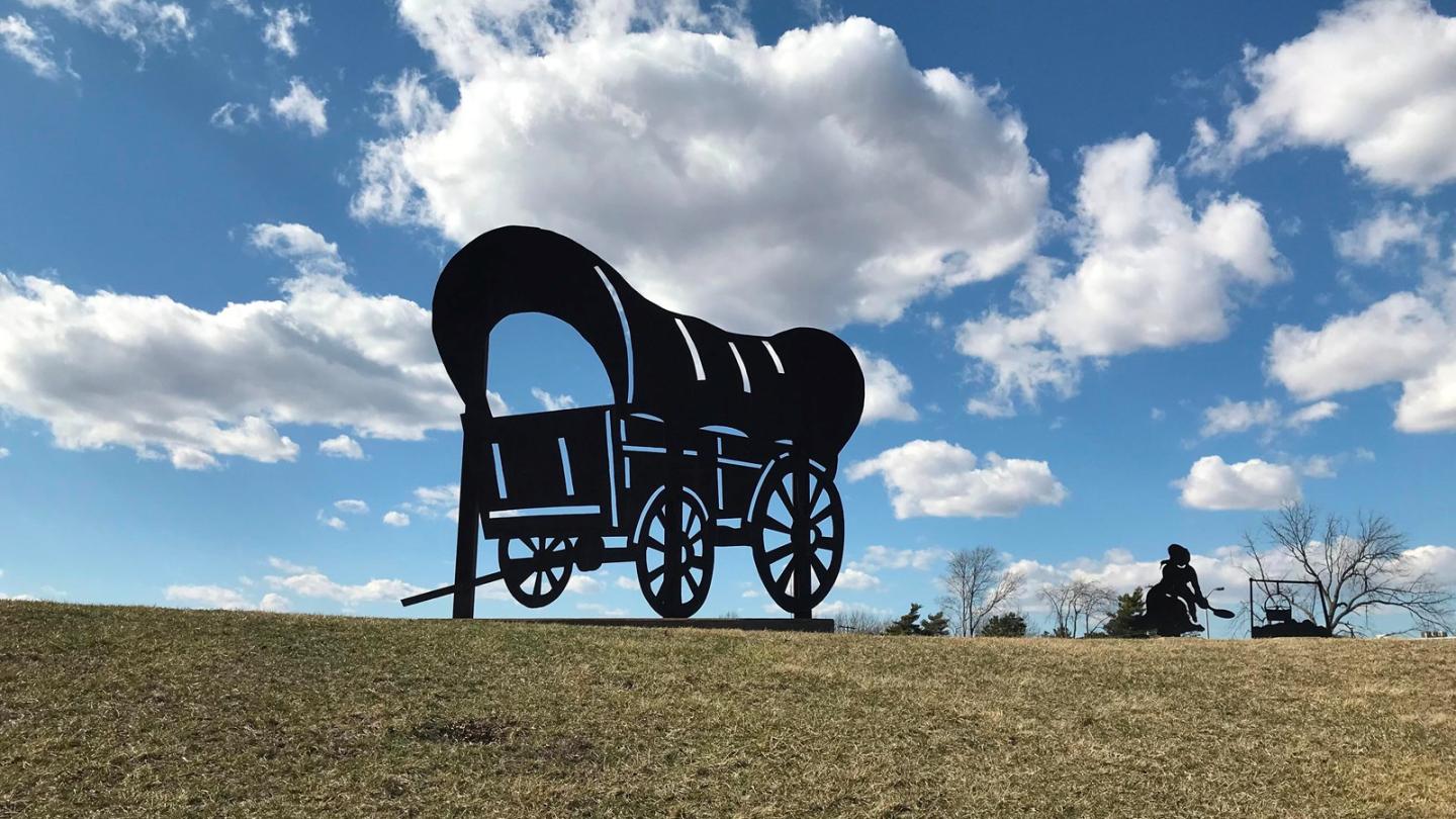 Silhouette of a covered wagon sculpture on a grassy hill under a cloudy blue sky.