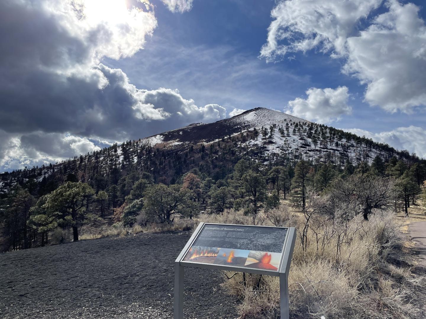 Mountain with trees, a cloudy sky, and snow patches; sign in foreground.
