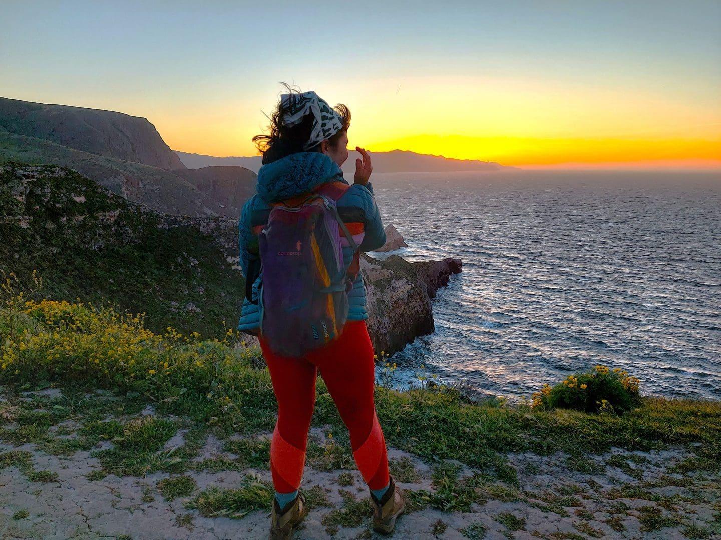 Hiker in colorful clothes watches ocean sunset from a cliff.
