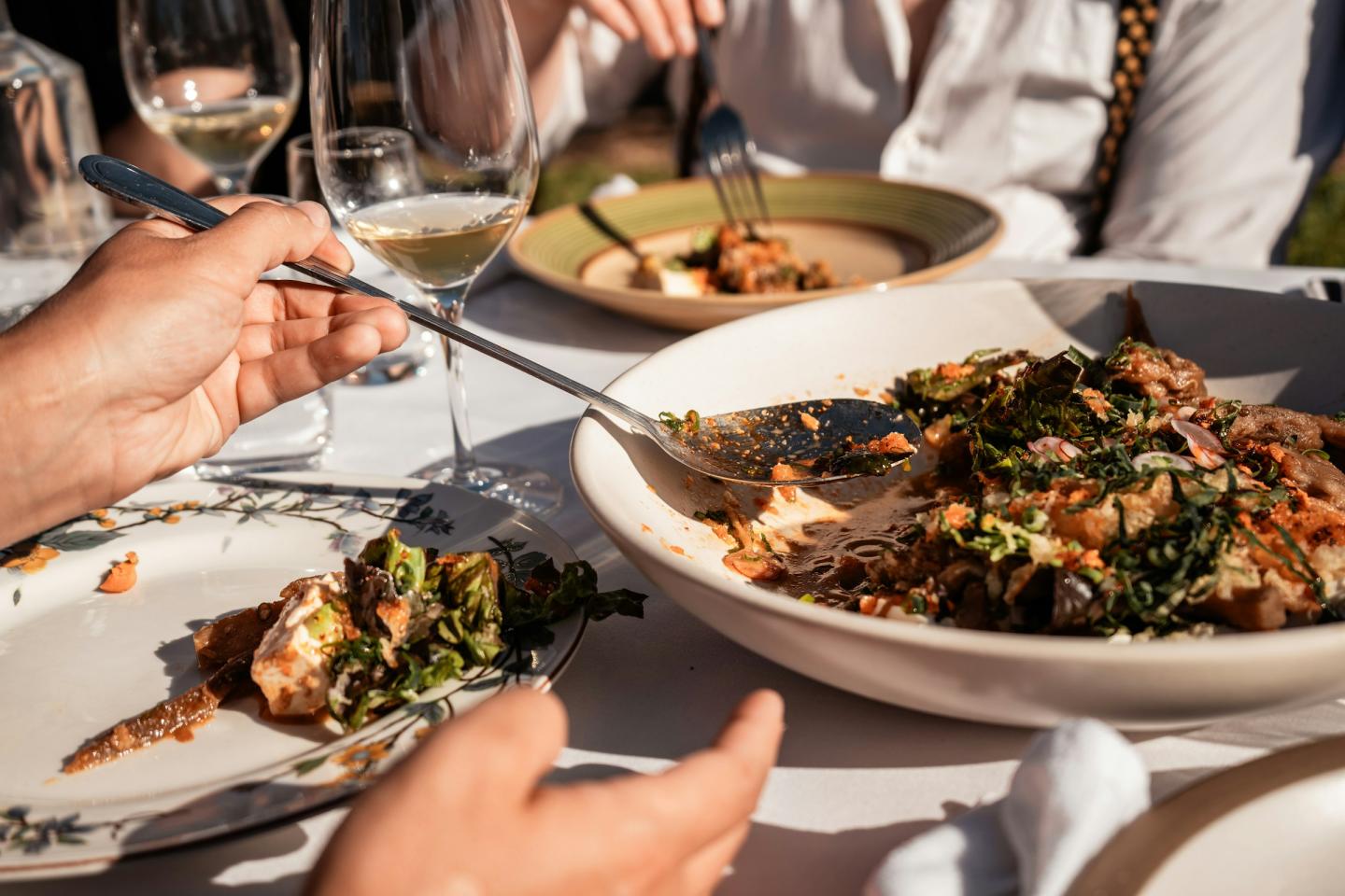 Hands serving salad at an outdoor dining table with wine glasses.
