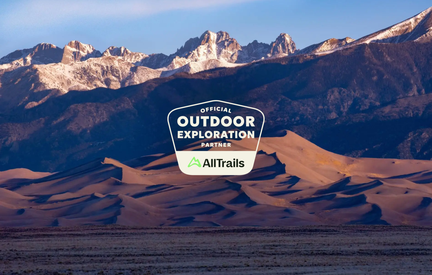 Sand dunes with snowy mountains in the background, AllTrails logo overlaid.