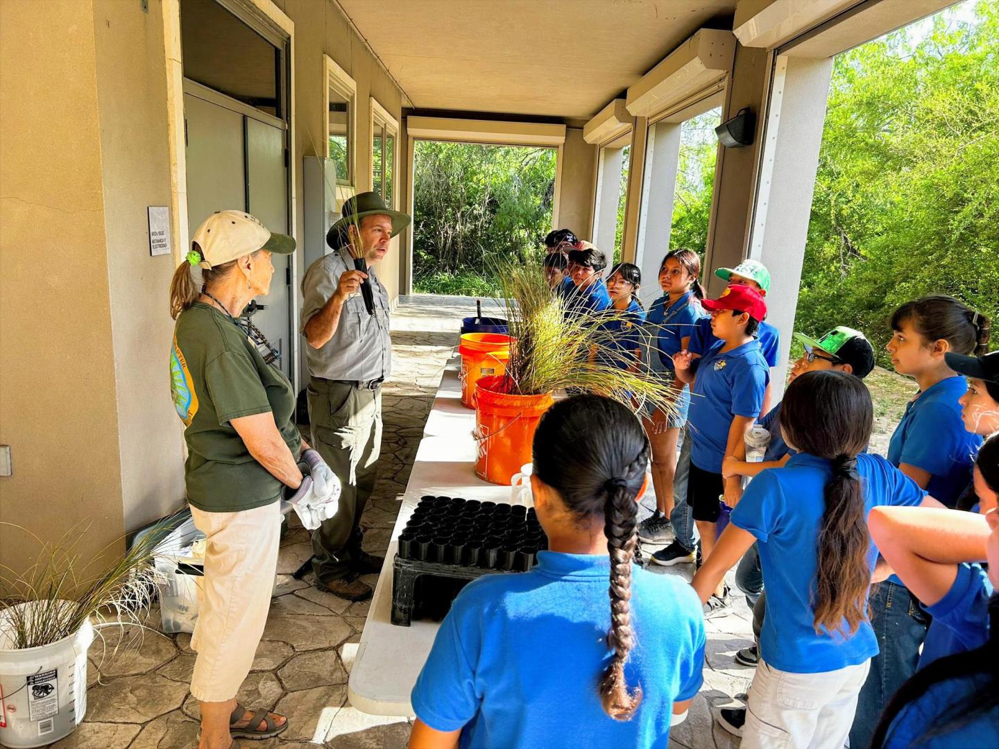 Outdoor class with an instructor and kids in blue shirts, surrounded by potted plants.