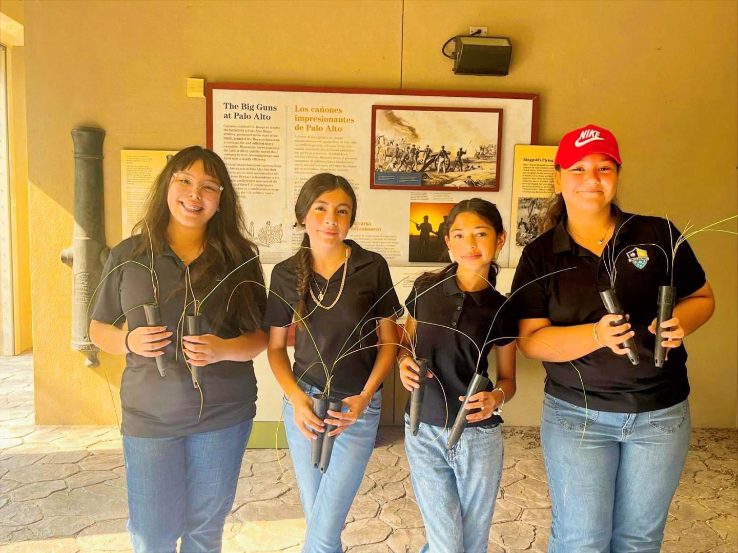 Four women smiling, holding audio equipment, wearing black shirts and jeans indoors.