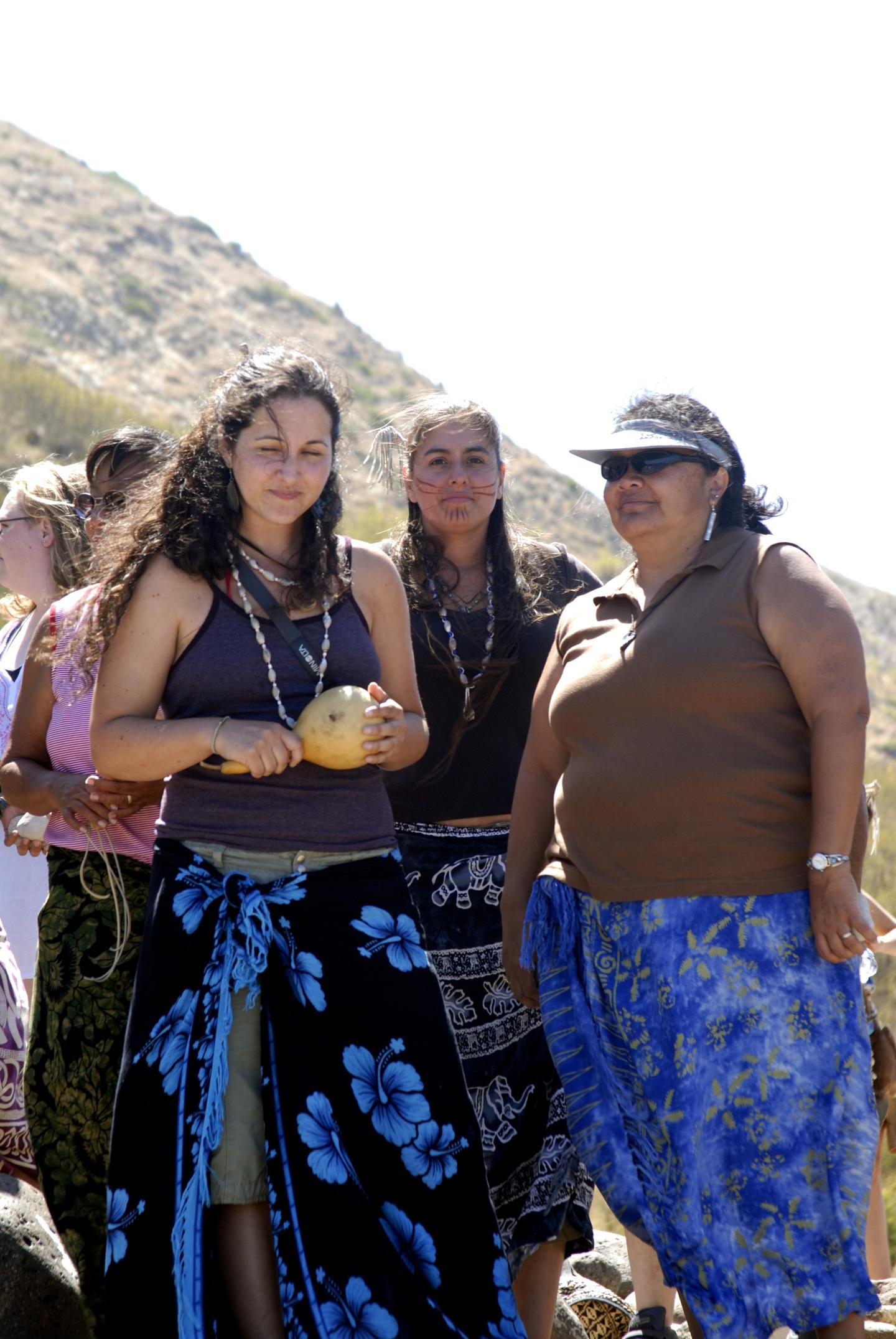 Women standing outdoors in sunny, hilly terrain, wearing colorful skirts.