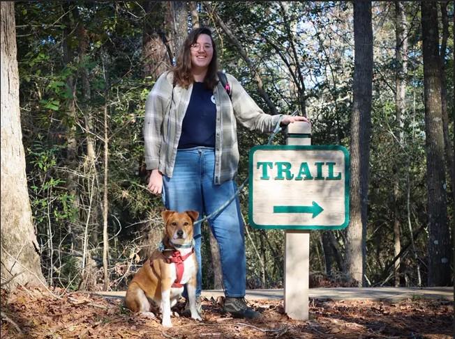 Woman with a dog standing by a trail sign in a wooded area.