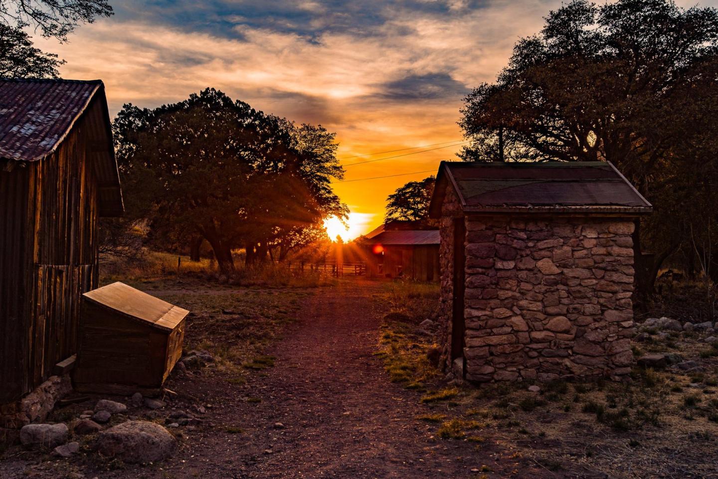 Sunset between rustic wooden and stone huts, with trees silhouetted against the colorful sky.