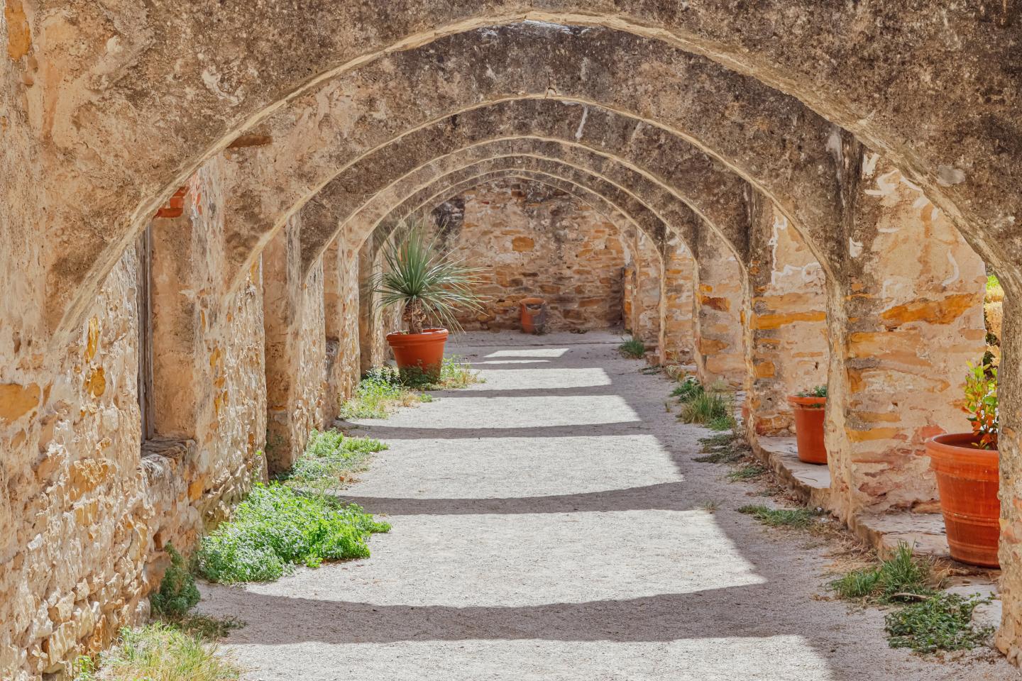 Stone arch corridor with red potted plants and sunlight casting shadows.