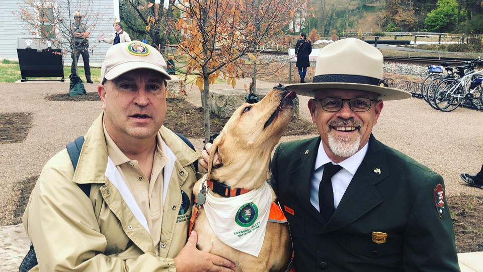 Two park rangers in uniform smile with a dog wearing a bandana.