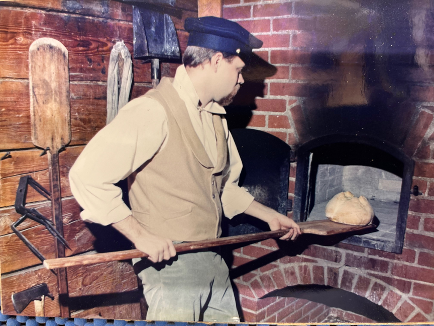 Baker in vintage attire using a wooden peel to place bread in brick oven.