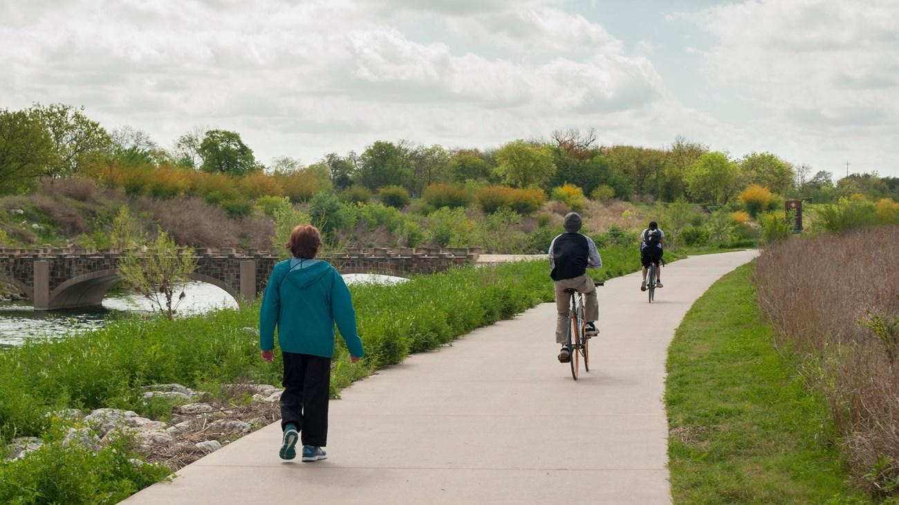 Pathway by river with people walking and biking, surrounded by greenery.