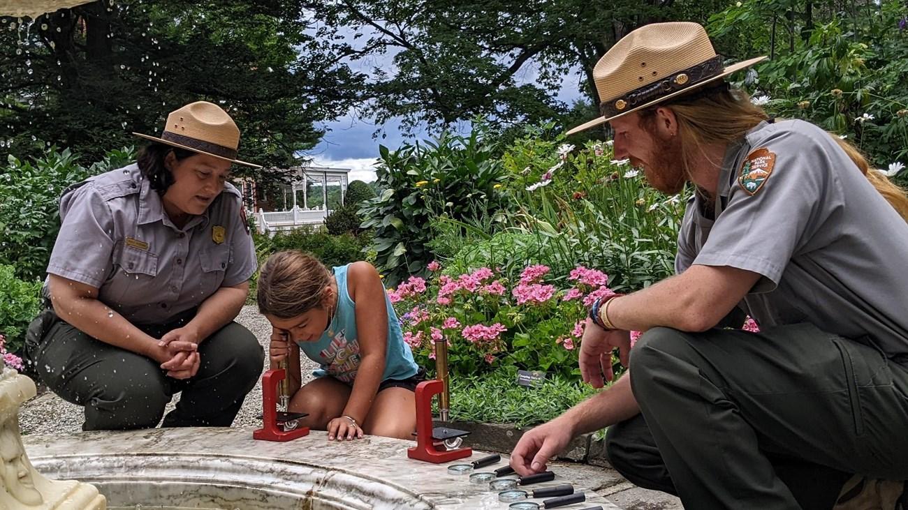 Child examines water with park rangers outside, surrounded by greenery and flowers.