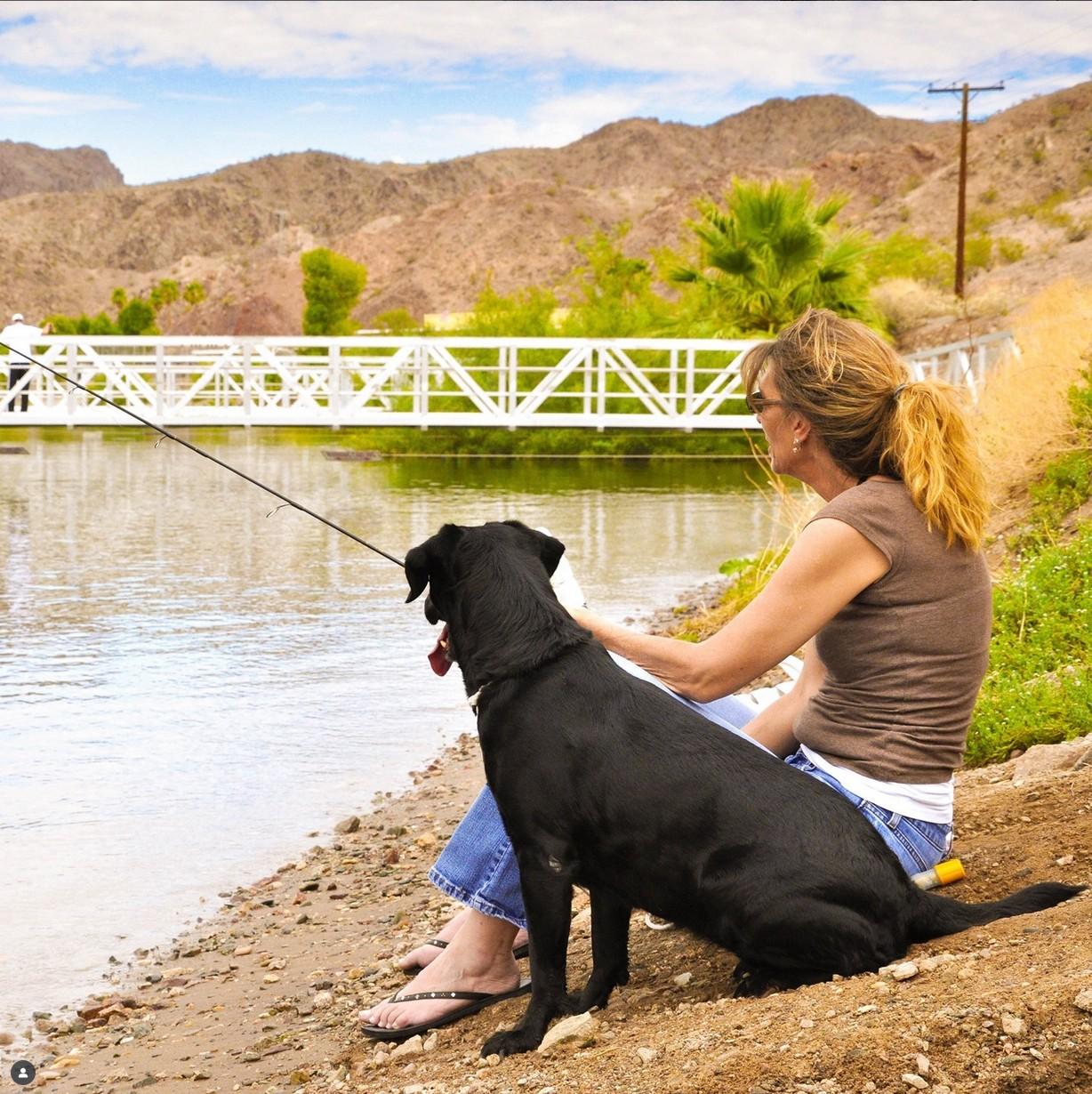 Woman and black dog sitting by a river, mountains in the background.