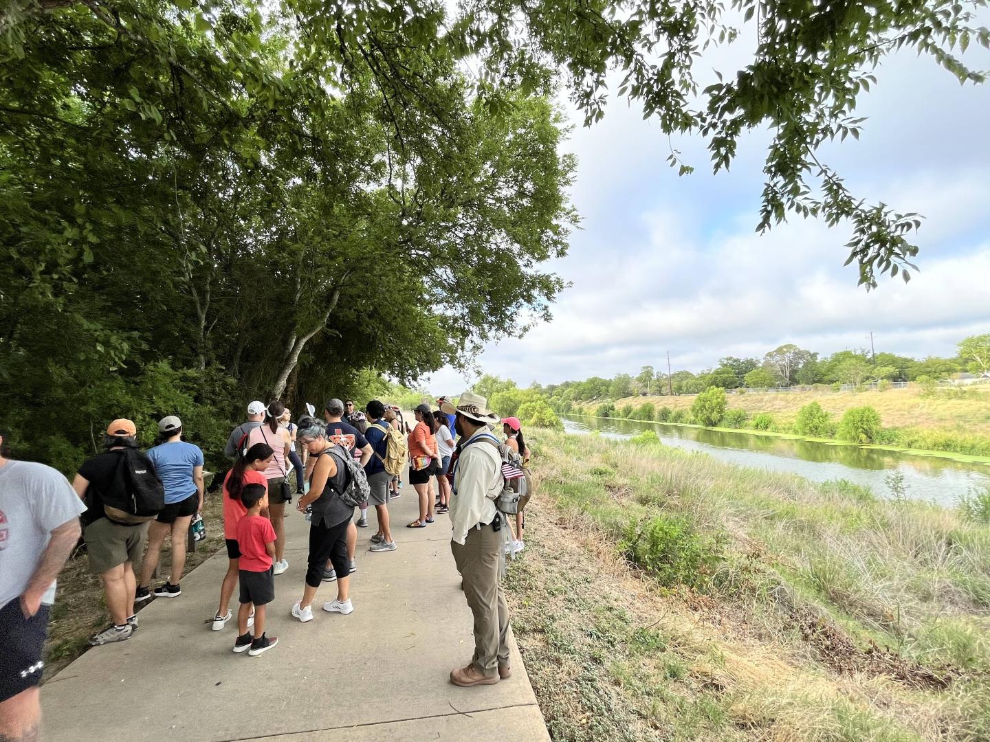 Group walking on a path by a tree-lined river under a blue sky.