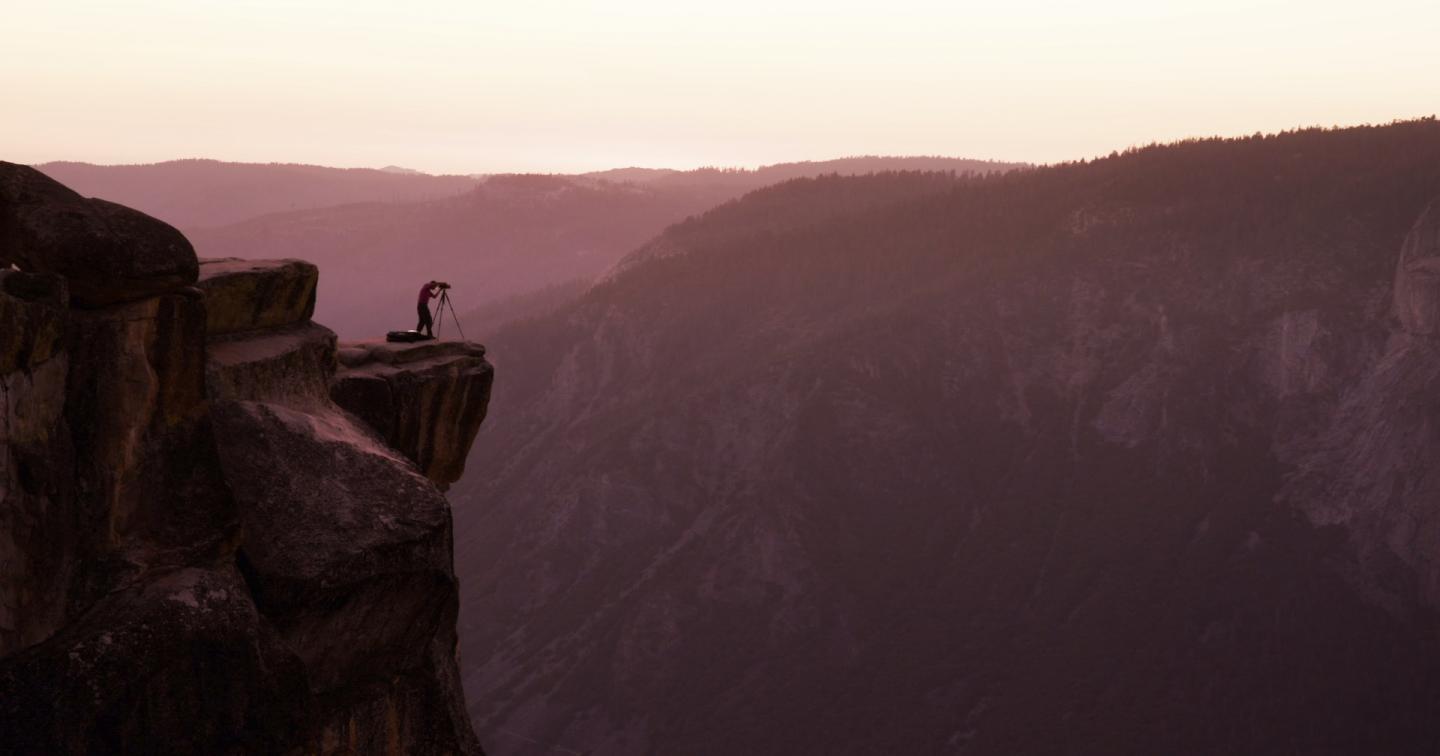 Person standing on a cliff edge at sunset, overlooking misty mountains.