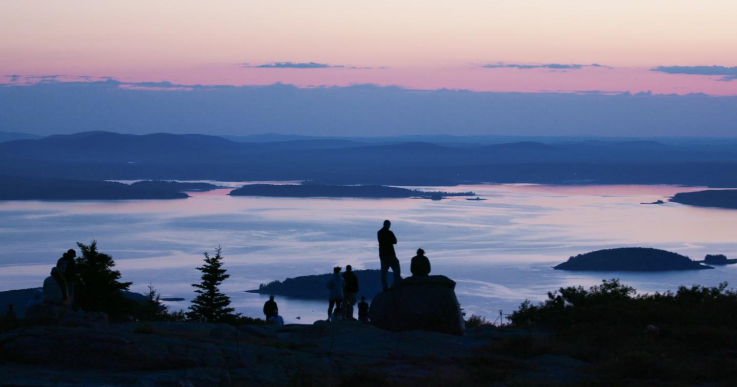 People silhouetted against a serene pink and blue sunset over a distant lake.