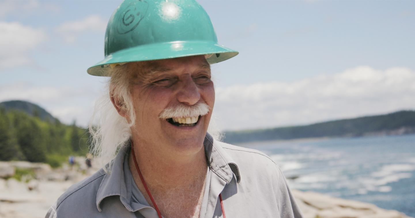 Smiling man in a green hard hat by the ocean on a sunny day.