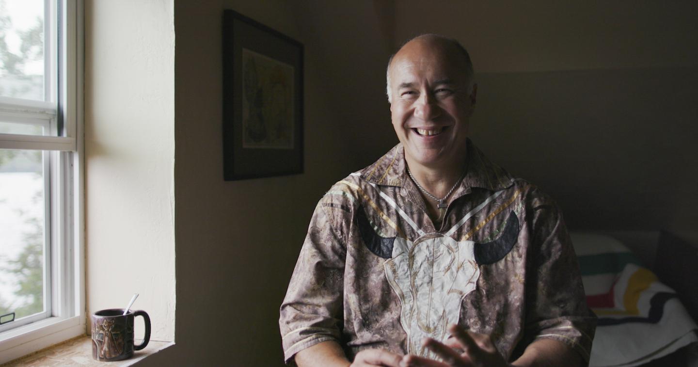 Smiling man sits near a window with a mug on the sill.