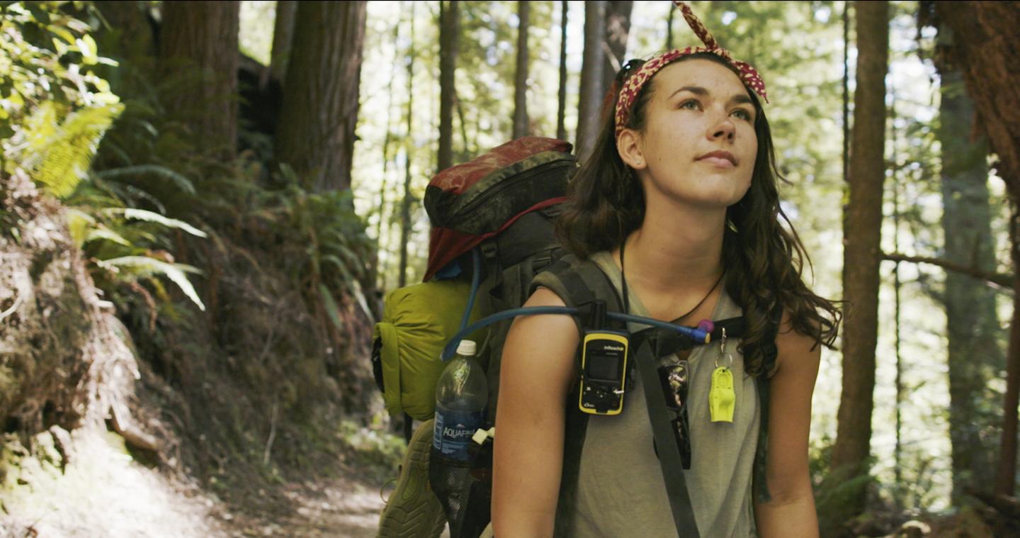 Woman hiking in a forest, wearing a backpack and bandana, looking upward.