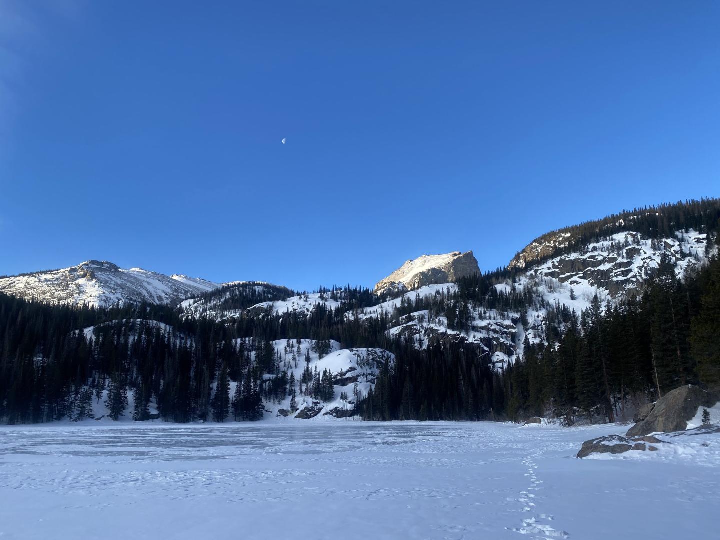 Snowy lake with mountains and trees under a blue sky.