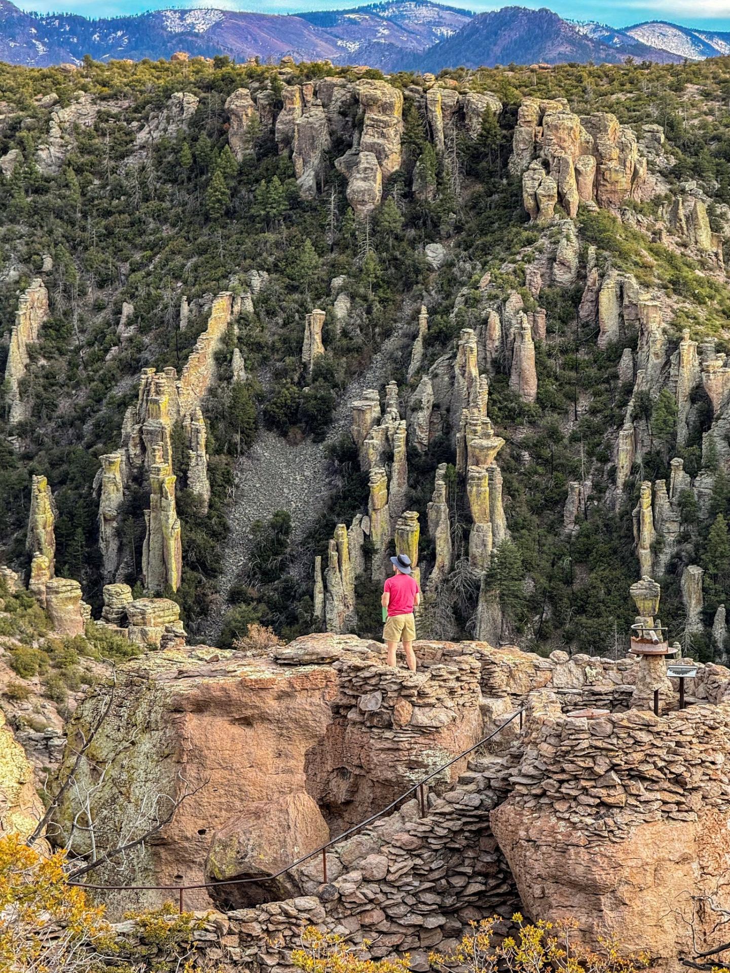 Man in red shirt stands on rocky ledge, overlooking tall rock formations and green hills.
