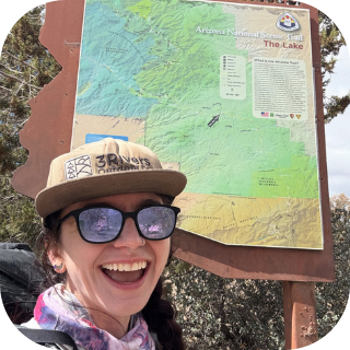 Smiling woman in sunglasses and hat by a colorful Arizona Trail map.