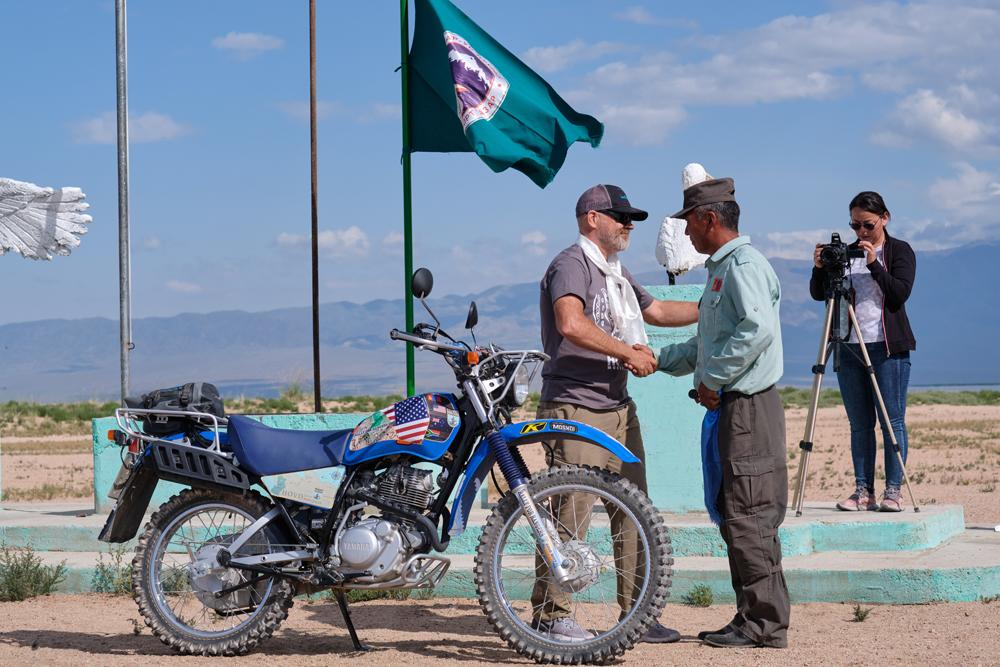 Two people shaking hands near a motorcycle, with flags and a photographer in the background.