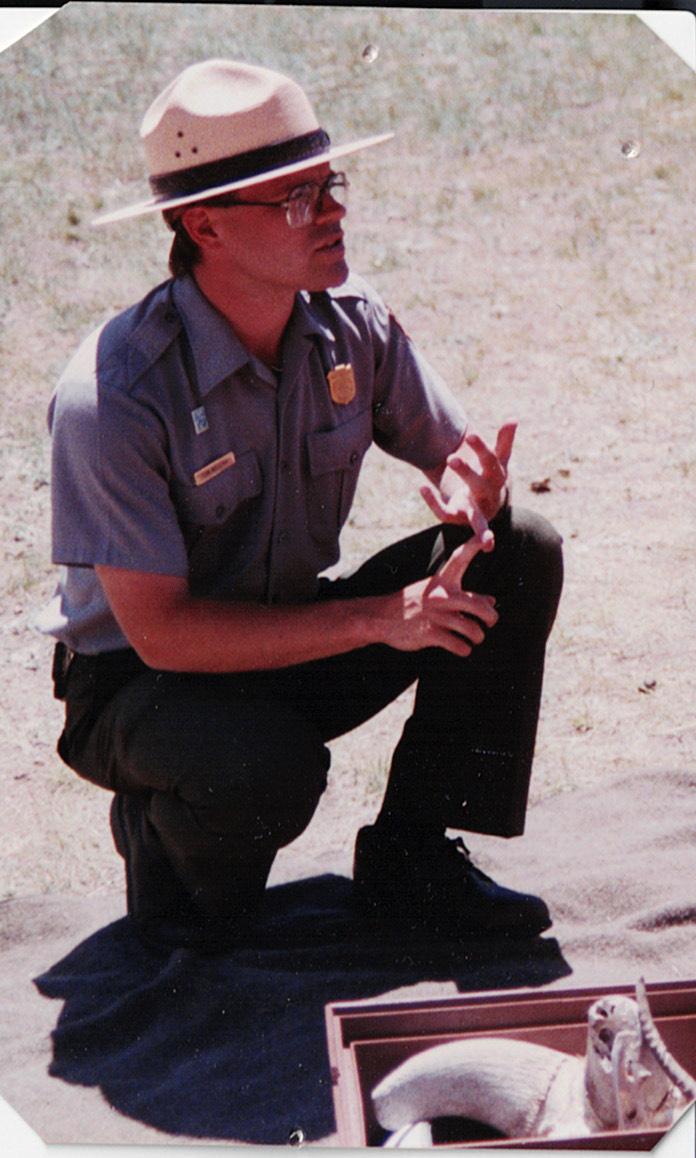 Park ranger in uniform kneels on the ground, talking and gesturing with hands.