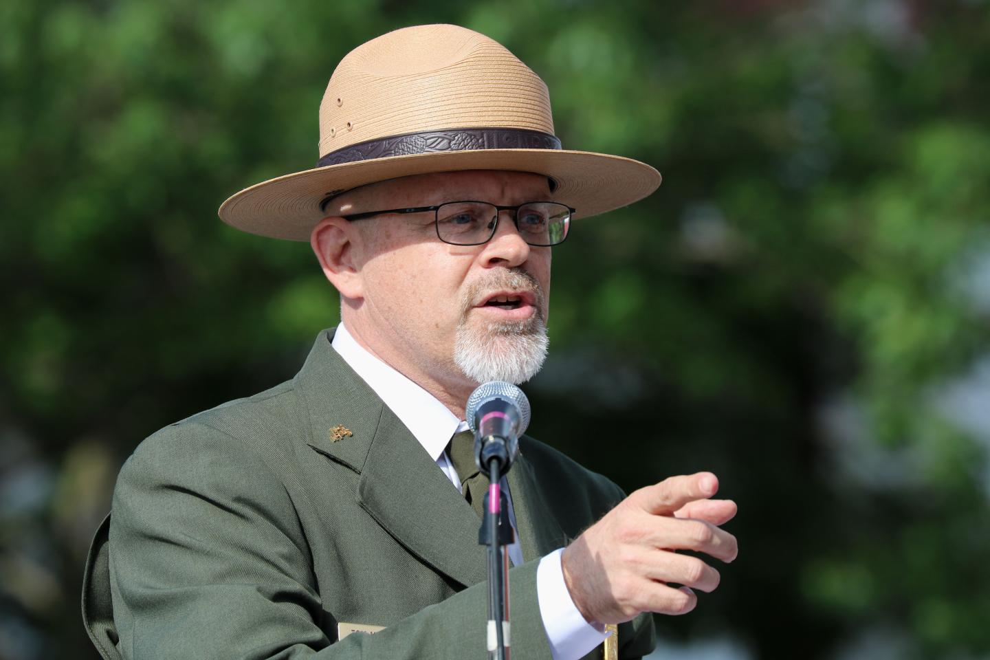 Man in ranger hat speaking into a microphone outdoors.