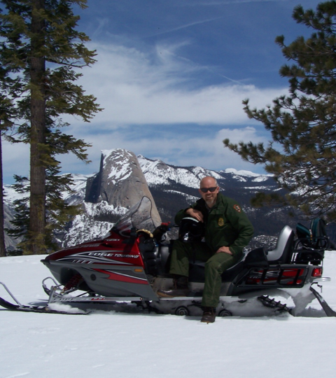 Man in uniform on a snowmobile, snowy mountain backdrop.