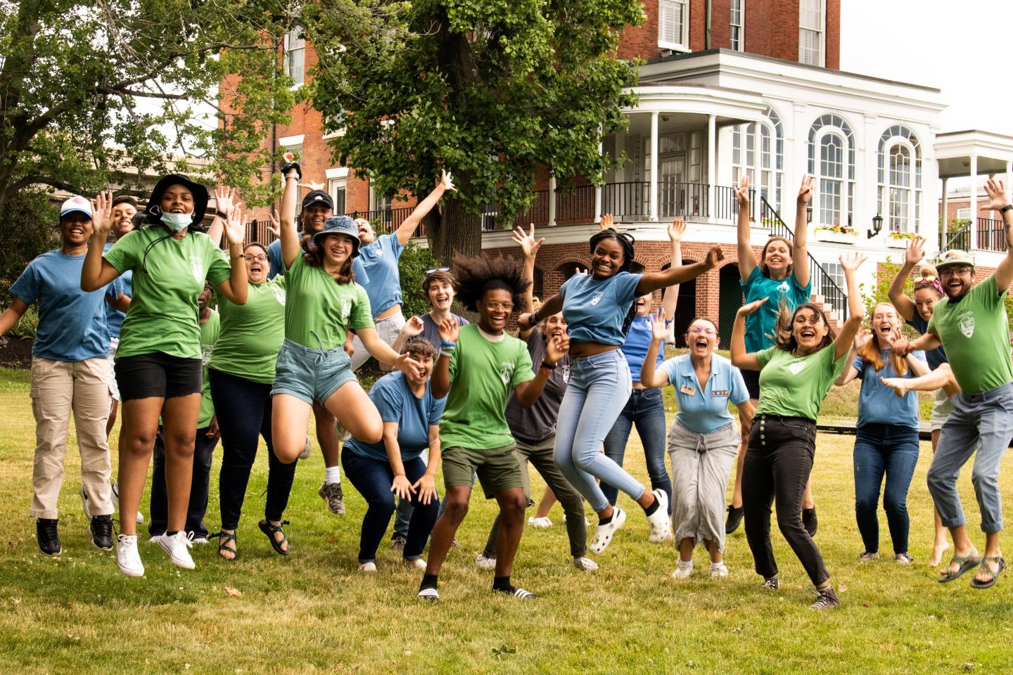 Group of people joyfully jumping on a green lawn, brick building in the background.