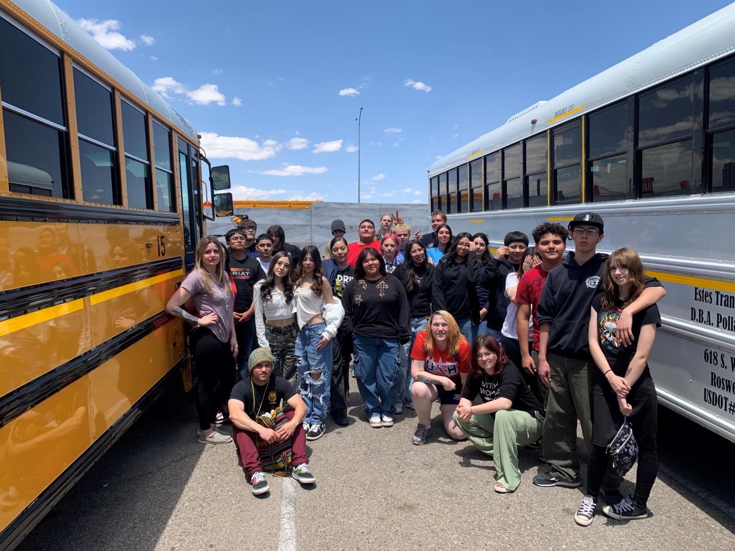 Group of people posing between two school buses in a sunny parking lot.