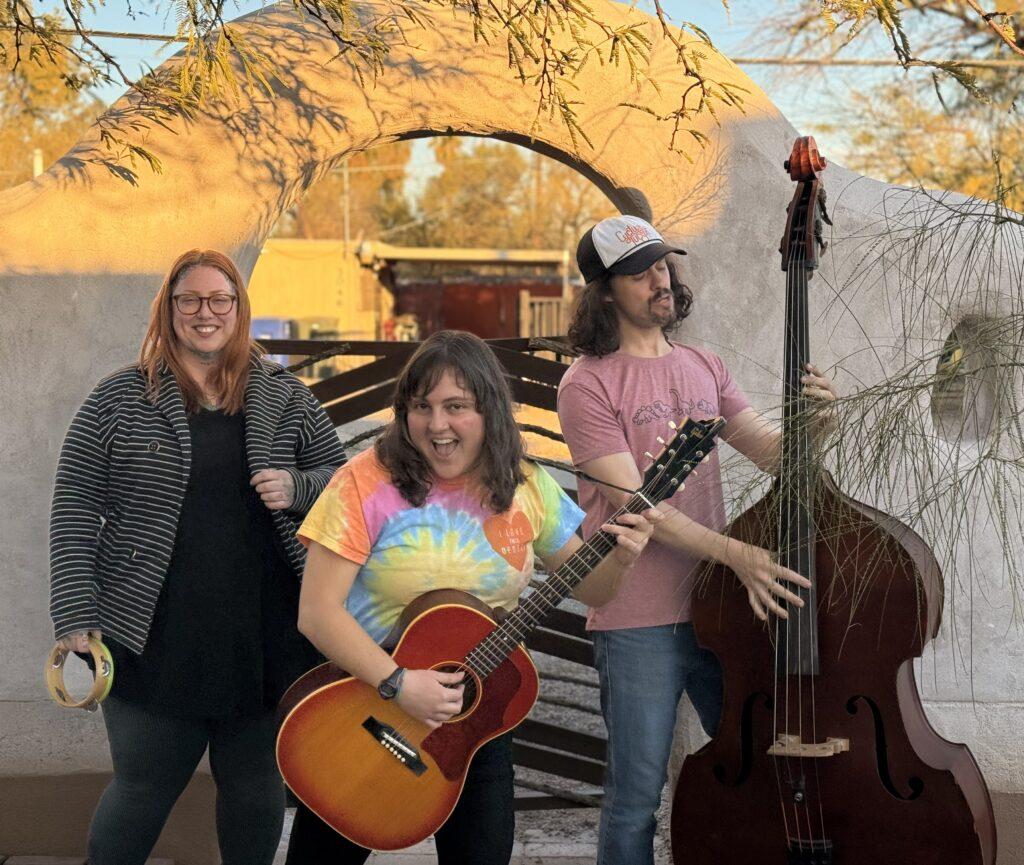 Three people play instruments outdoors, one with a tambourine, guitar, and upright bass.