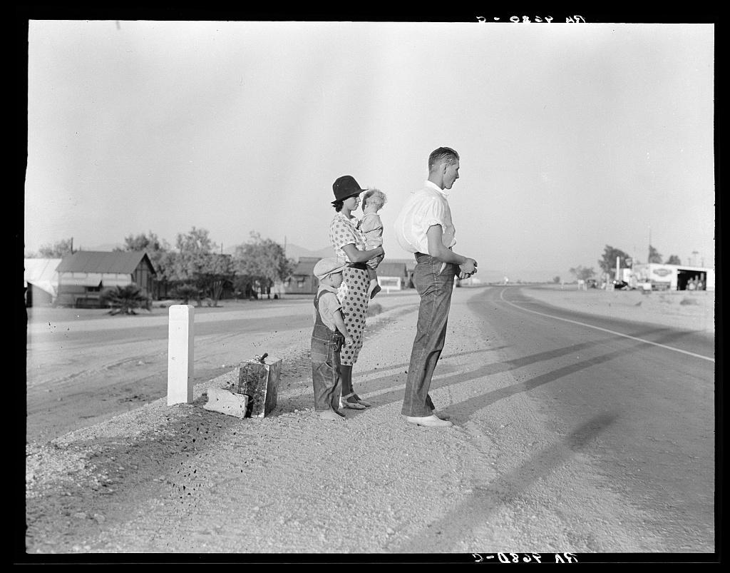 Man, woman, and child standing by a roadside, with a suitcase and distant houses.