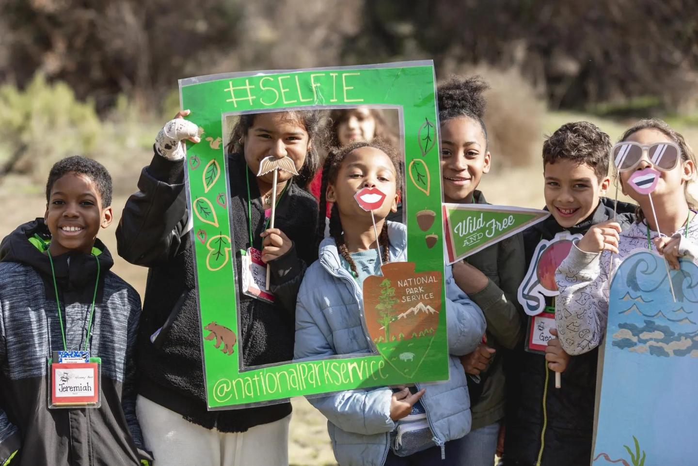 Group of 7 young people standing together and smiling, holding photo booth props and posing with a large, green, rectangular frame that reads #selfie and "National Parks" Service.