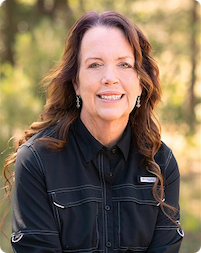 Smiling woman outdoors in a black shirt, surrounded by trees.