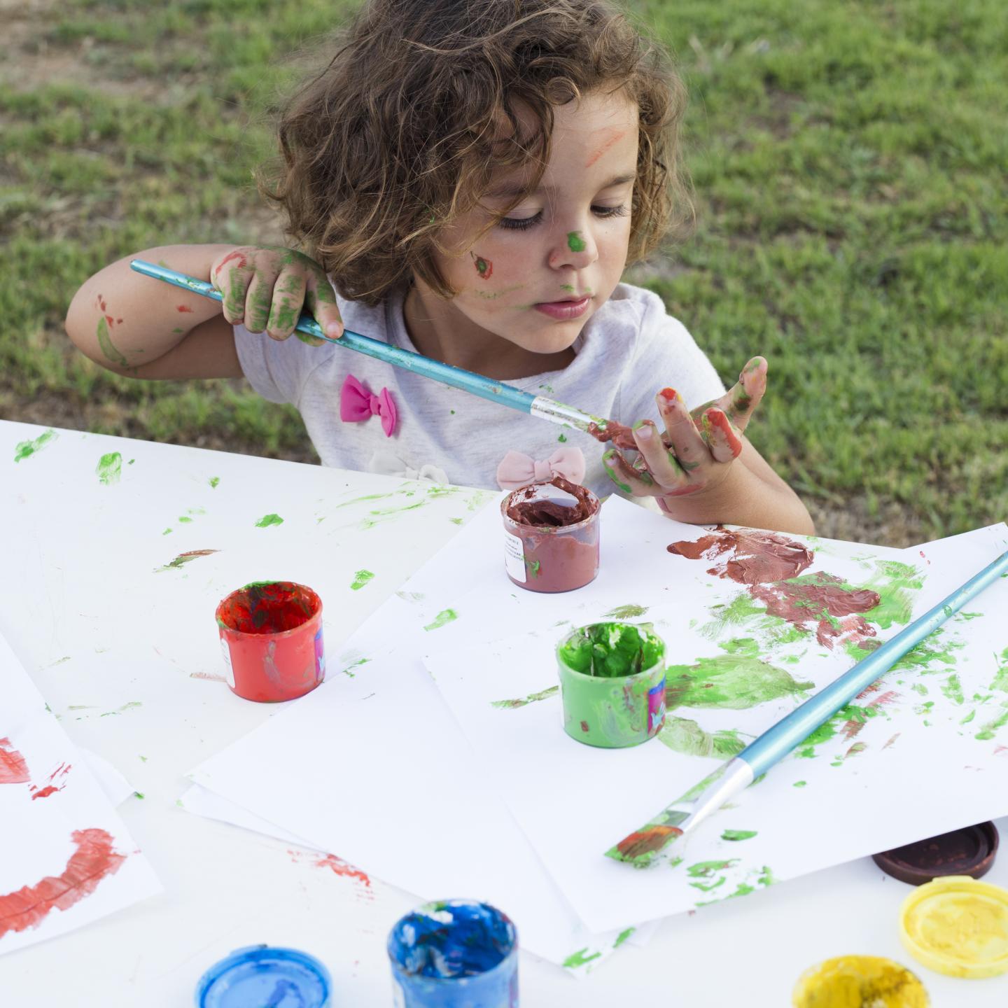 Child painting with colorful paints outdoors at a table.