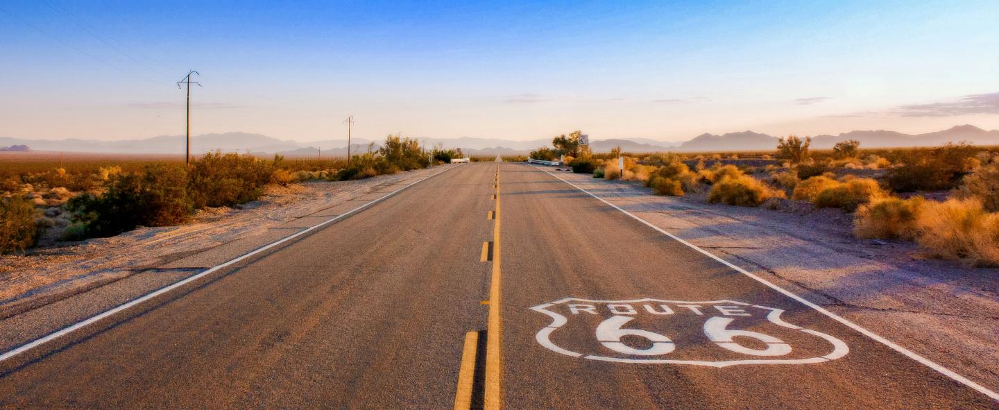 Desert highway with Route 66 logo, blue sky, and distant mountains.