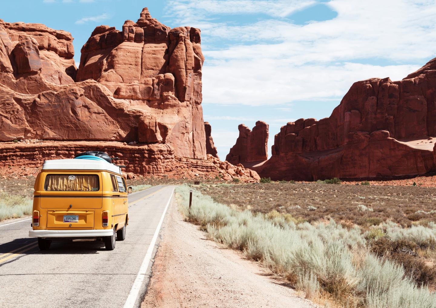 Yellow van driving through desert landscape with red rock formations.