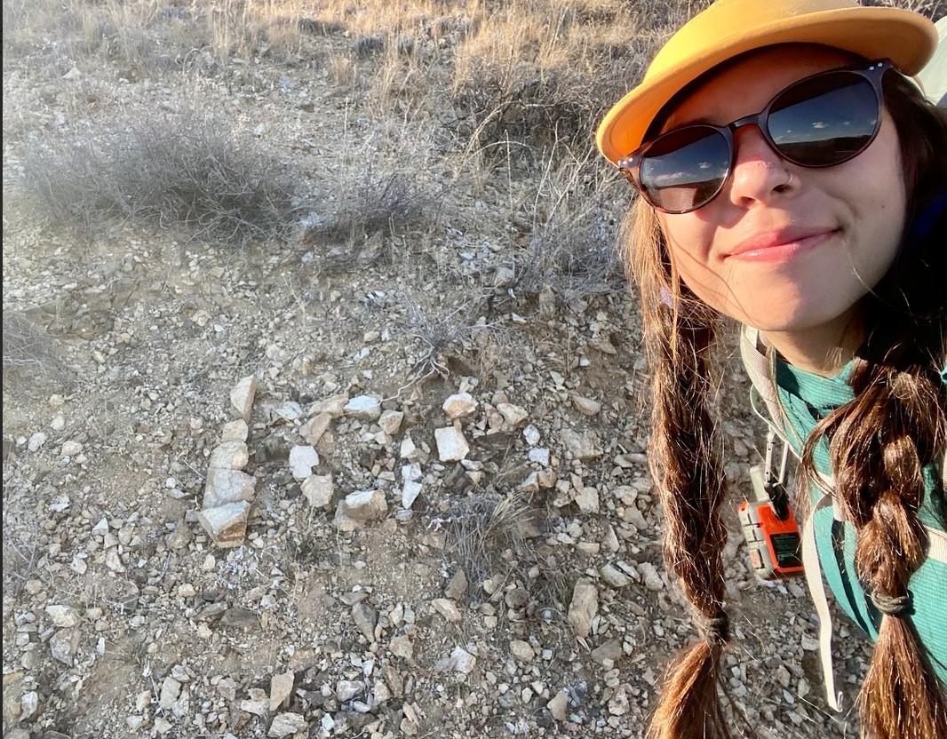 Smiling person wearing a yellow cap, standing on rocky ground.